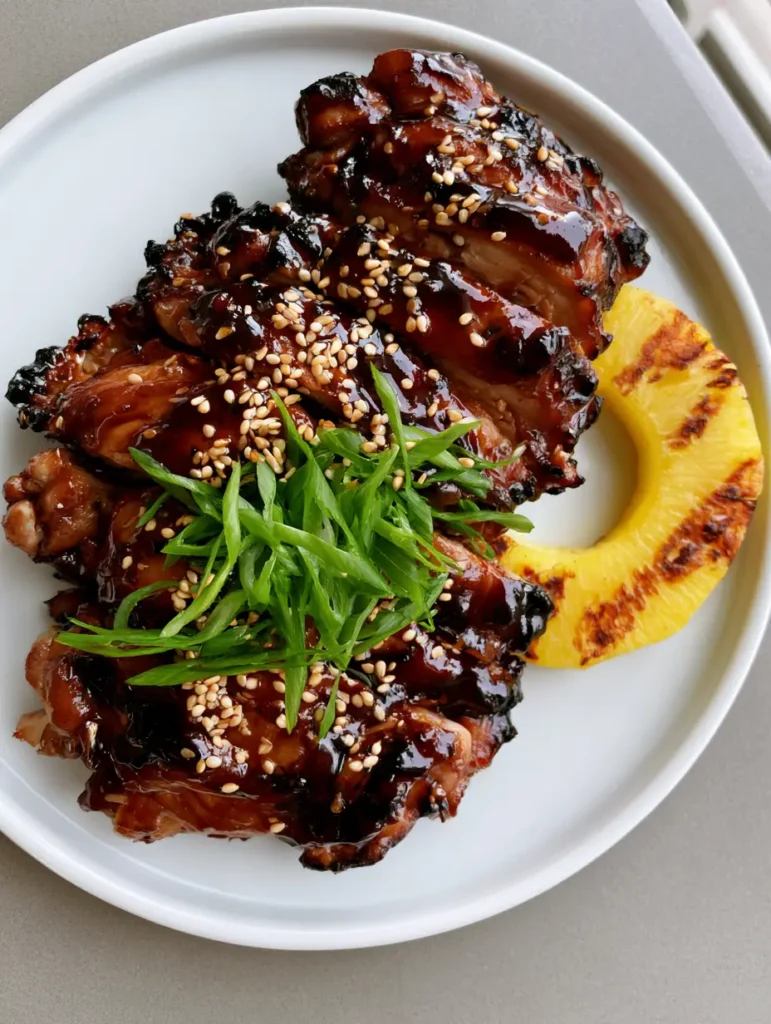 Slices of teriyaki chicken garnished with bright green scallions and sesame seeds next to charred yellow pineapple.