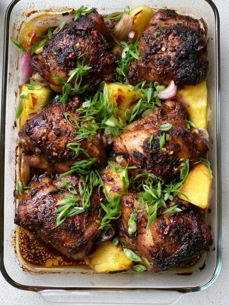 Overhead view of a glass baking dish with Pineapple Jerk Chicken, translucent red onions, and golden roasted pineapple chunks.
