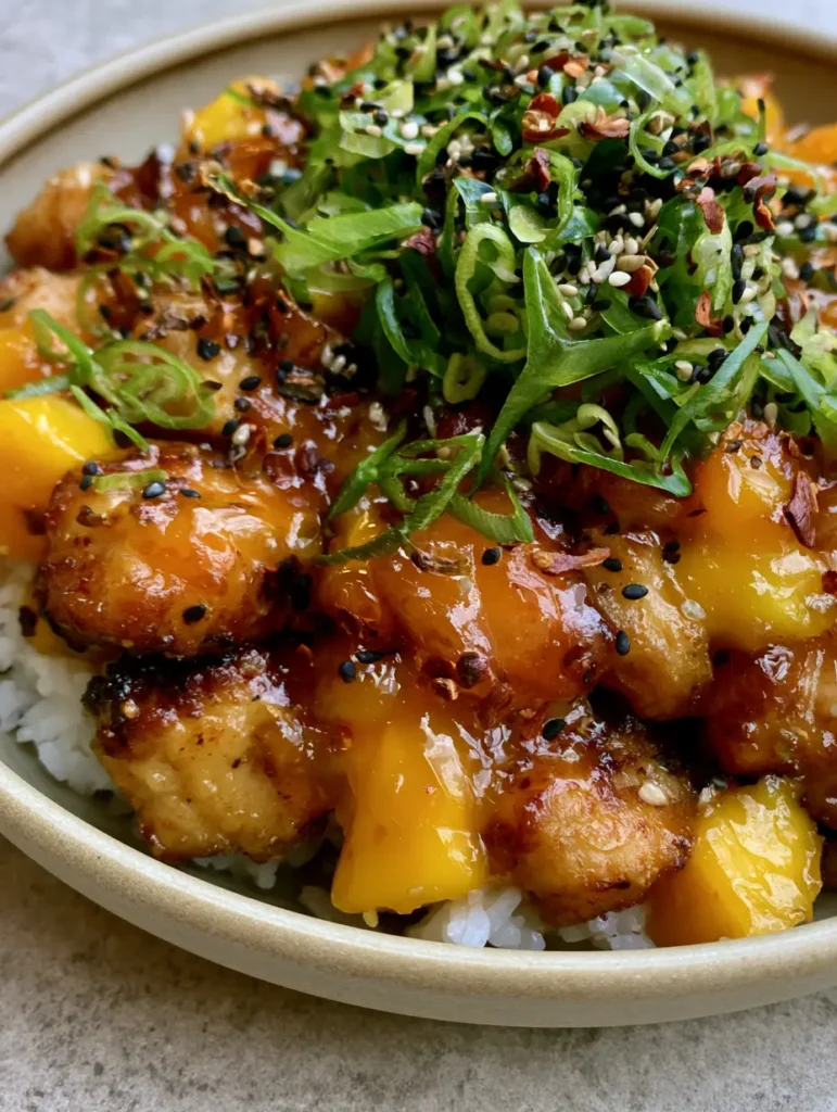 Bowl of Mango Glazed Chicken with fresh mango chunks and heavy sesame seed garnish on a grey stone countertop.